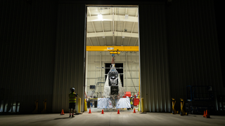 NASA Goddard astrophysicist Kyle Helson looks at EXCITE (EXoplanet Climate Infrared TElescope) as it dangles from the ceiling of a hangar at NASA’s Columbia Scientific Balloon Facility in Fort Sumner, New Mexico.

Credit: NASA/Sophia Roberts

Alt text: A man looks at a large telescope in a hangar. 

Image description: A crane suspends a shiny silver telescope in a large hangar at night. The top is conical, with a section cut out for a cylinder. The body is rhombus-shaped and has two shiny rectangular panels attached to the bottom that extend slightly in front of the telescope. The background shows the hangar is full of equipment, and the foreground shows the outside of the building. There are orange cones in front of the hanger doors. A person in a reflective vest and hard hat stands to the left of the open doors.