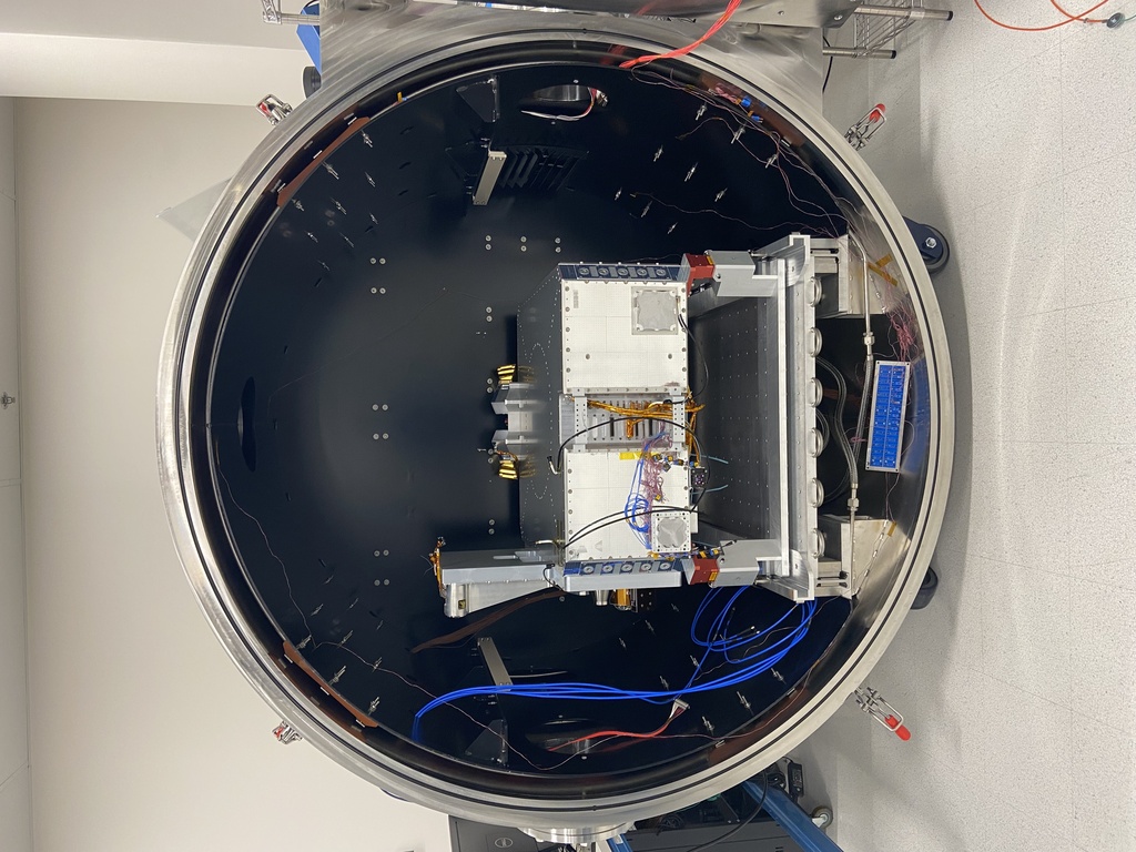 Pandora’s spacecraft bus was photographed Jan. 10 within a thermal-vacuum testing chamber at Blue Canyon Technologies in Lafayette, Colorado. The bus provides the structure, power, and other systems that will enable the mission to help astronomers better separate stellar features from the spectra of transiting planets.Credit: NASA/Weston Maughan, BCTImage description: In this vertical photo, a metal box placed on a short stand with red supports rests within a large, dark cylindrical chamber. A bundle of blue wires extends from the upper left of the chamber into a coil at the bottom left of the stand. Blue, black, pink, and gold wires can be seen connecting to the front of the box. The background is an off-white wall, and a column is visible at right.